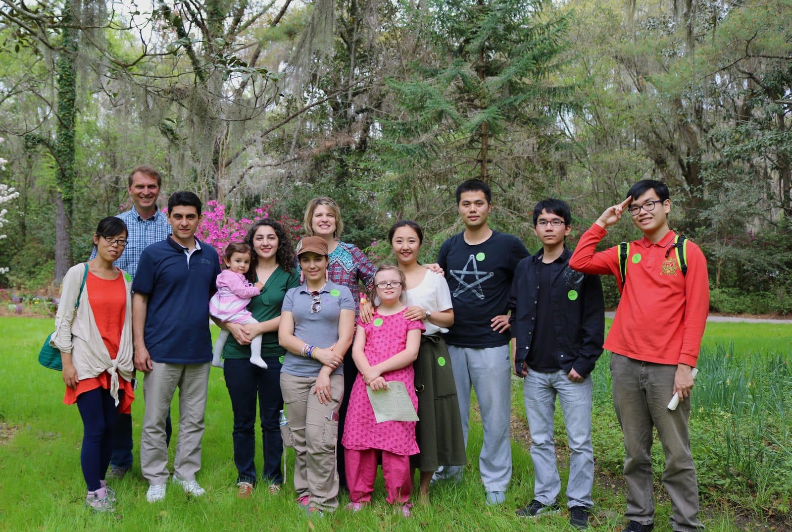 About a dozen students and friends of all ages, including a toddler held by her mother and a girl in a pink dress, stand together on a lawn under moss-hung trees; most wear green stickers and the young man at the right salutes.