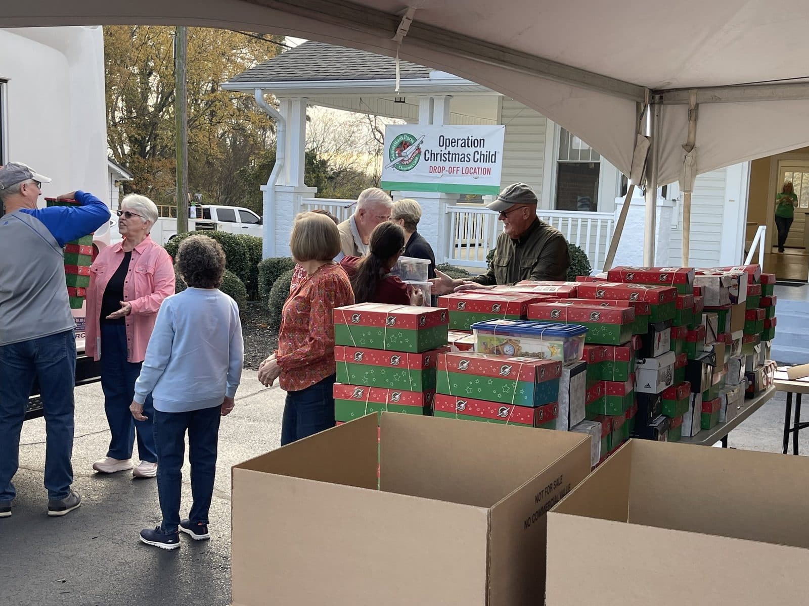 Volunteers sort a wall of shoeboxes under the tent in front of the drop-off location banner.